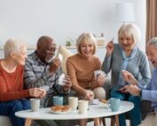 Diverse seniors sitting together on a couch, laughing and playing a card game, highlighting social activities in a senior living community.